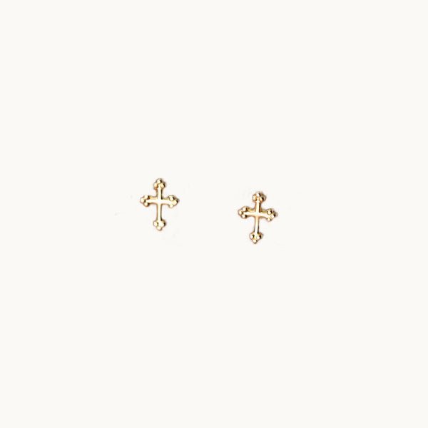 Une paire de boucles d'oreilles est exposée devant un fond beige.  Ce sont des petites croix avec trois points au bout de chaque extrémité. ce sont des boucles en or.