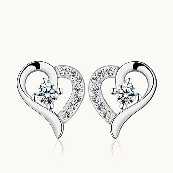 Une paire de boucles d'oreilles est exposée devant un fond beige.  Ce sont des boucles d'oreille en forme de coeur. Une partie du coeur est en argent et l'autre côté est en strass. Il y a un diamant au milieu du coeur. Elles existent en argent 925. 