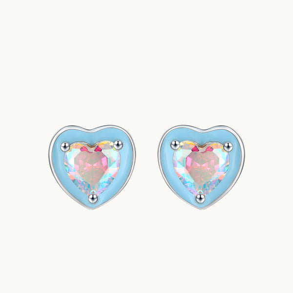 Une paire de boucles d'oreilles est exposée devant un fond beige.  Ce sont des boucles en forme de coeur bleu ornées au milieu d'un coeur en diamant de zirconium. Le diamant est bleu et rose ce qui donne de très jolis reflets. Elles sont en argent 925. 