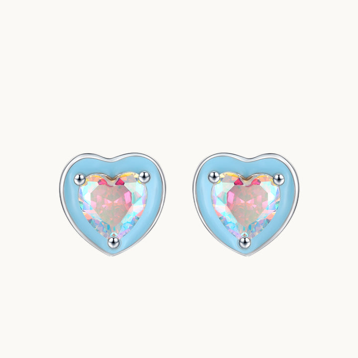 Une paire de boucles d'oreilles est exposée devant un fond beige.  Ce sont des boucles en forme de coeur bleu ornées au milieu d'un coeur en diamant de zirconium. Le diamant est bleu et rose ce qui donne de très jolis reflets. Elles sont en argent 925. 