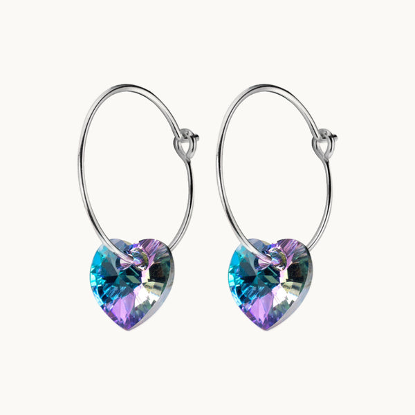 Une paire de boucles d'oreilles est exposée devant un fond beige.  Ce sont des créoles fines sur lesquelles pend un cœur en strass violet et bleu. Elles existent en argent 925.