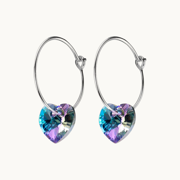 Une paire de boucles d'oreilles est exposée devant un fond beige.  Ce sont des créoles fines sur lesquelles pend un cœur en strass violet et bleu. Elles existent en argent 925.