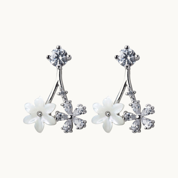 Une paire de boucles d'oreilles est exposée devant un fond beige.  Ce sont des boucles d'oreille constituées d'un strass sur lequel pend une fleur blanche et une fleur avec des pétales en diamant.  Elles sont en argent 925.