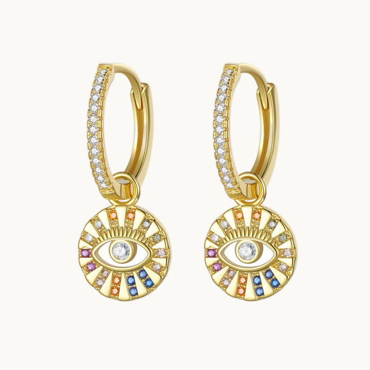 Une paire de boucles d'oreilles est exposée devant un fond beige.  Ce sont des anneaux en strass desquels pendent un rond en strass multicolores formant un oeil.  Elles sont en argent 925 plaqué or.