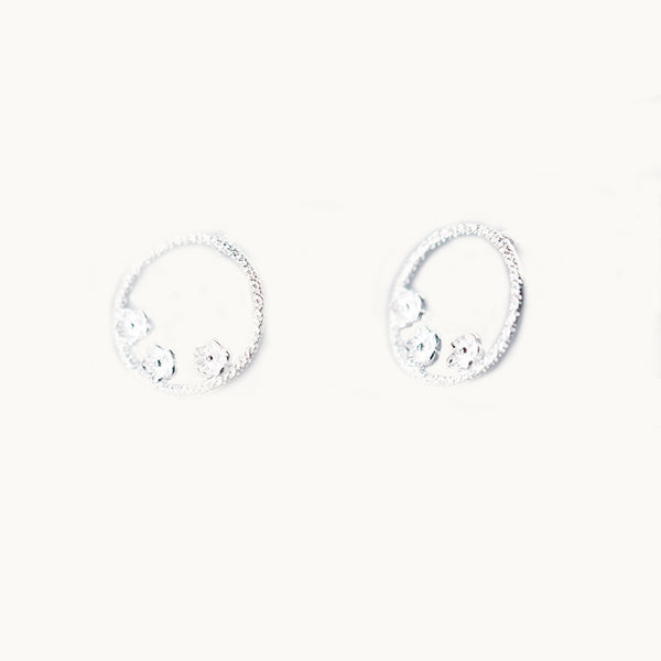 Une paire de boucles d'oreilles est exposée devant un fond beige.   Ce sont des boucles d'oreille ronde en argent 925 ornées de 3 petites fleurs. 