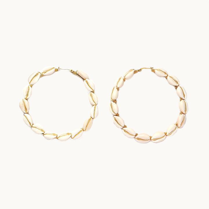  Une paire de boucles d'oreilles est exposée devant un fond beige.  Ce sont des boucles d'oreilles créoles en plaqué or ornées de coquillages 