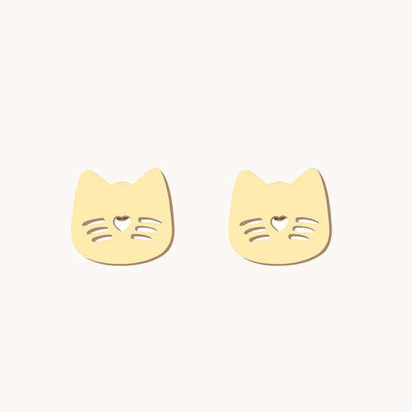 Une paire de boucles d'oreilles est exposée devant un fond beige.  Elles sont en forme de chat avec moustache et museau. Elles sont dorées. 