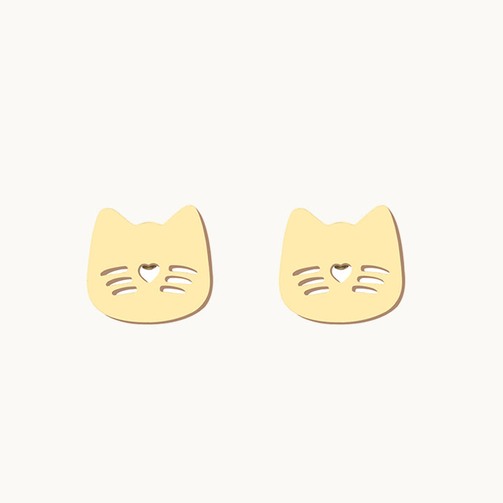 Une paire de boucles d'oreilles est exposée devant un fond beige.  Elles sont en forme de chat avec moustache et museau. Elles sont dorées. 