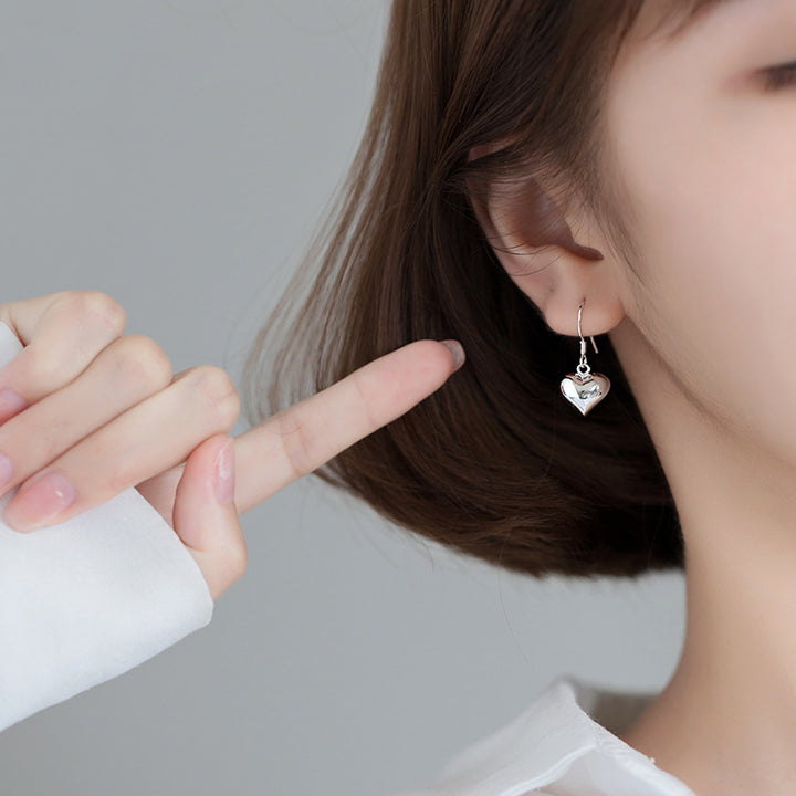 Une femme pointant son oreille avec des boucles d'oreilles pendantes en forme de cœur en argent 925. Un bijou élégant pour tous les jours ou des occasions spéciales.