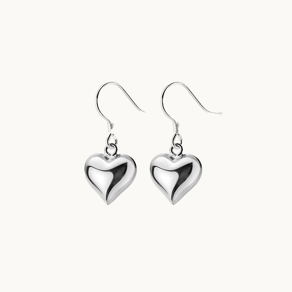 Une paire de boucles d'oreilles est exposée devant un fond beige.  Ce sont des boucles d'oreilles pendantes composées d'un coeur argent 925. 