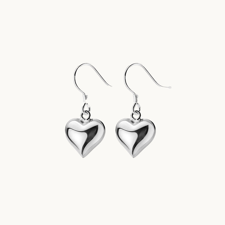 Une paire de boucles d'oreilles est exposée devant un fond beige.  Ce sont des boucles d'oreilles pendantes composées d'un coeur argent 925. 