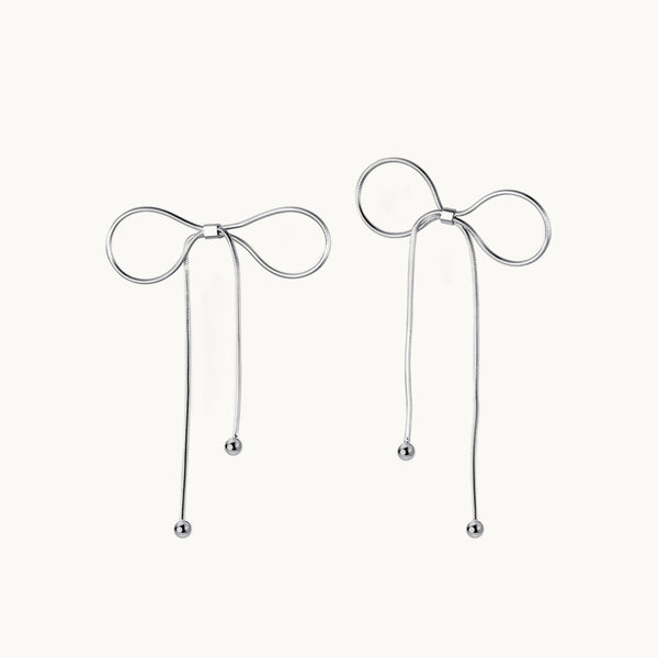 Une paire de boucles d'oreilles est exposée devant un fond beige.   Ce sont des boucles en forme de noeud fin. Elles sont en argent 925.