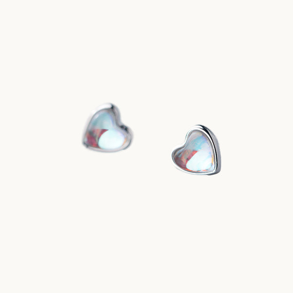 Une paire de boucles d'oreilles est exposée devant un fond beige.  Ce sont des boucles en forme de coeur composées en opale. L'opale est clair avec des reflets arc-en-ciel. Elles sont en argent 925.