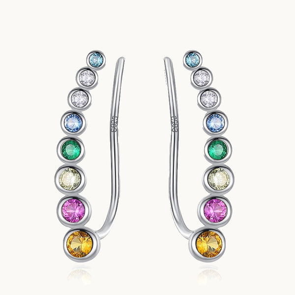 Une paire de boucles d'oreilles est exposée devant un fond beige.  Ce sont des boucles grimpantes constituées de strass alignés du plus grand au plus petit. Elles sont  en argent 925 avec des strass multicolores.