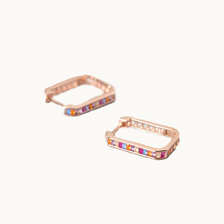 Une paire de boucles d'oreilles est exposée devant un fond beige.  Ce sont des créoles à la forme rectangulaire. Elles sont ornées de strass colorés. Elles sont en argent 925 plaqué or rose. 