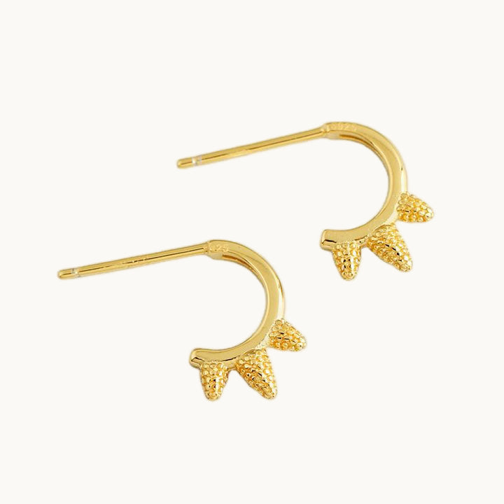 Une paire de boucles d'oreilles est exposée devant un fond beige.  Ce sont des boucles anneaux avec 3 piques sur chacune. Ce sont des boucles en or. 