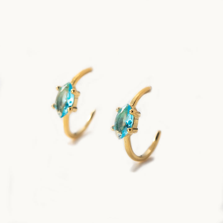 Une paire de boucles d'oreilles est exposée devant un fond beige.  Ce sont des boucles d'oreille en forme d'anneau incomplet qui est ornée sur le devant d'un diamant bleu ciel en forme d'amande, appelé "marquise". Elles sont en argent 925 plaqué or.