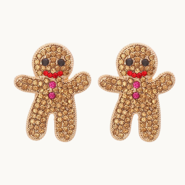 Une paire de boucles d'oreille est exposée devant un fond beige.  Ce sont des boucles d'oreille tout en strass en forme de bonhomme de pain d'épice. Elles sont idéales pour les fêtes de noël. Elles sont dorées. 