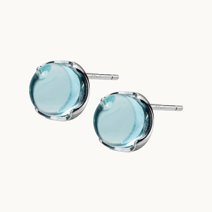 Une paire de boucles d'oreilles est exposée devant un fond beige.  Ce sont des boucles rondes en cristal bleu ciel. Elles sont en argent 925.