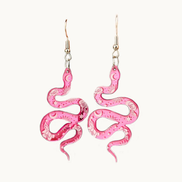 Une paire de boucles d'oreilles est exposée devant un fond beige.  Ce sont des boucles d'oreille pendante. L'attache est argenté. De cette attache pend un serpent de couleur rose sur lequel sont dessinées des lunes, des fleurs et des étoiles. Le serpent est fabriqué en résine.