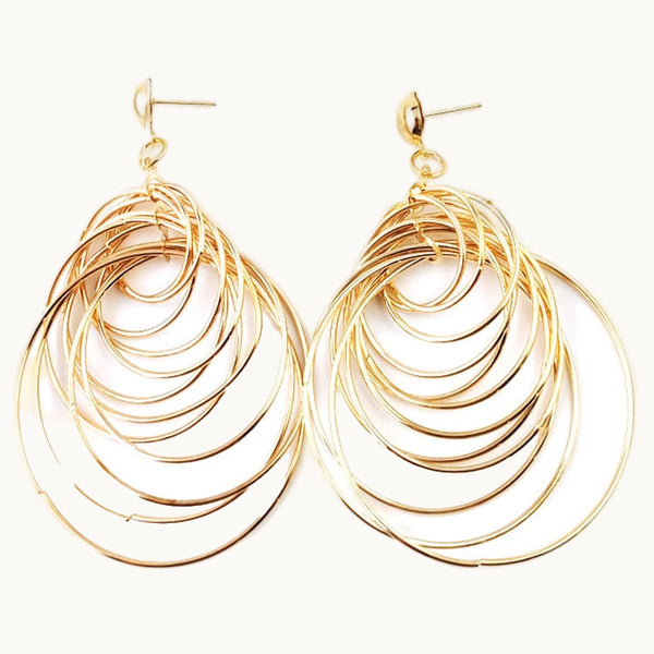 Une paire de boucles d'oreille est exposée devant un fond beige.  Ce sont des boucles d'oreille dorées pendantes composées de multiples anneaux entrelacés.