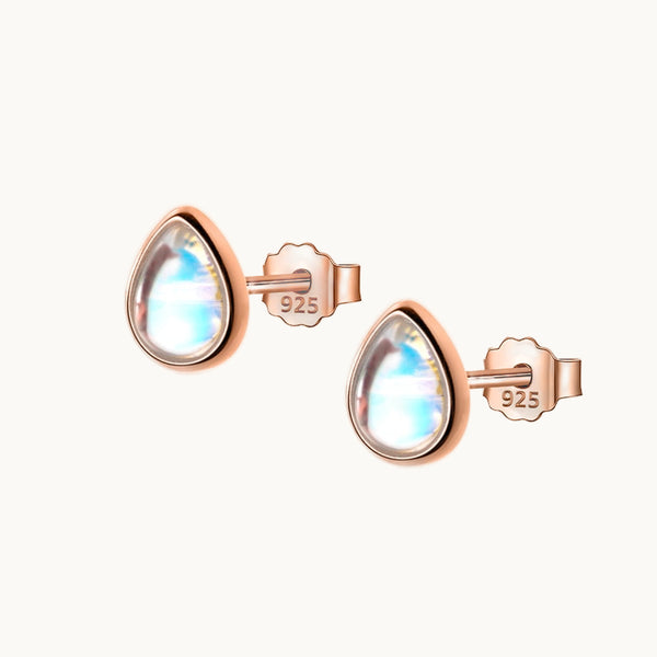 Une paire de boucles d'oreilles est exposée devant un fond beige.  Ce sont des boucles d'oreille qui ressemblent à de véritables gouttes d'eau. Elles ont une pierre transparente bleutée avec des reflets qui rappellent une goutte d'eau au soleil. La forme de la boucle est une goutte. Elles sont en plaqué or rose. 