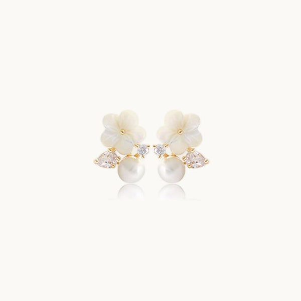 Une paire de boucles d'oreilles est exposée devant un fond beige.  Ce sont de petites boucles d'oreille délicates. Elles sont composées d'une fleur blanche, d'une perle et de strass (un rond et un en amande.) Elles sont en argent 925 plaqué or.
