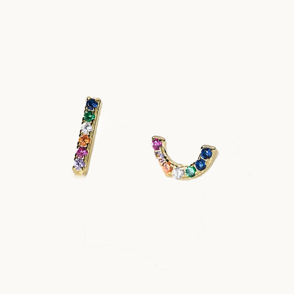 Une paire de boucles d'oreilles est exposée devant un fond beige.  Ce sont des boucles dépareillées. Il y a une boucle rectangulaire et une en arc de cercle. Elles sont toutes les deux ornées de strass multicolores. Les boucles sont en or. 