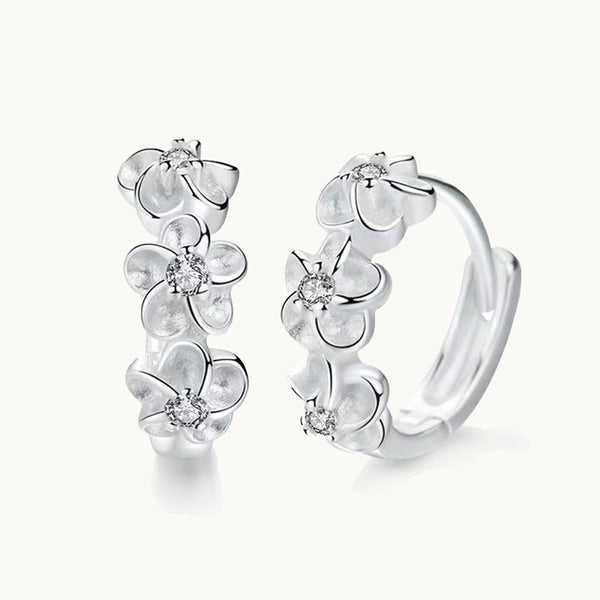 Une paire de boucles d'oreilles est exposée devant un fond beige.  Ce sont des créoles ornées de trois fleurs. Le coeur des fleurs est un petit strass. Elles sont en argent 925.