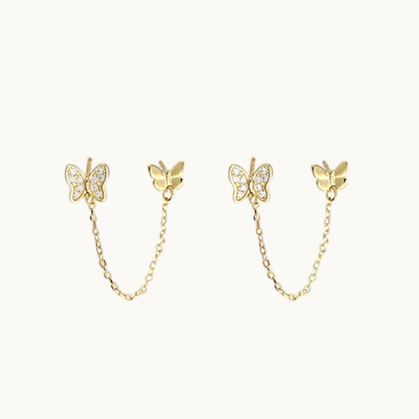 Une paire de boucles d'oreilles est exposée devant un fond beige.  Ce sont deux papillons dorées, l'un avec strass l'autre sans avec une chaîne qui les relie entre eux. 