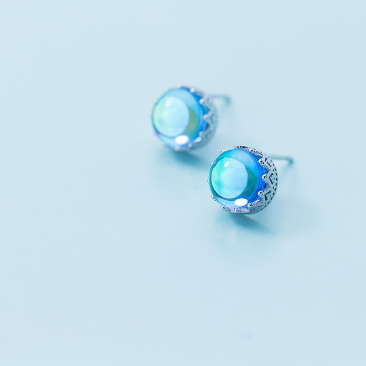 Une paire de boucles d'oreilles en forme de couronne avec un cristal bleu rond irradie des reflets arc-en-ciel, entouré d'argent 925. Bijou d'exception pour chaque occasion. Dimensions : 1 x 1 cm.