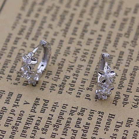 Boucles d'oreilles créoles fleurs strass argent 925 sur journal. Élégance inégalée avec 3 fleurs scintillantes ornées de strass étincelants en leur cœur. Fusion de simplicité et d'extravagance pour une touche d'éclat et de féminité. Poids : 1,09 g. Dimensions : 14 x 12 mm. Genre : Femme. Style : Boucle d'oreille strass.