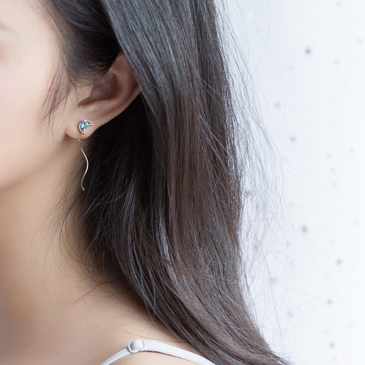 Une femme aux cheveux longs portant une boucle d'oreille en argent avec une étoile et une lune. Boucle d'oreille pendante dépareillée avec diamant de zirconium bleu saphir. Style élégant et original pour femme.