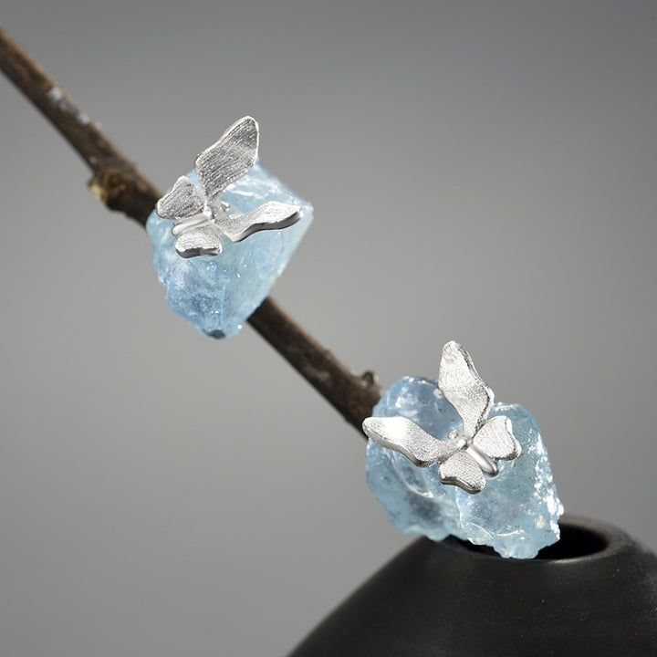Une paire de boucles d'oreilles est exposée devant un fond beige.  Ce sont des pierres bleues sur lesquelles sont posées des papillons en argent 925.
