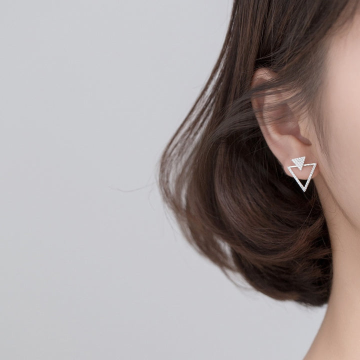 Une femme porte une boucle d'oreille double triangles strass en argent 925. Le triangle plein étincelant est délicatement serti de strass, surmontant un triangle ajouré plus grand, éclatant avec des strass sur deux de ses côtés. Un jeu visuel séduisant pour une touche de raffinement.