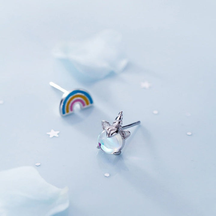 Une paire de boucles d'oreille dépareillées en argent 925 représentant un arc-en-ciel coloré et une licorne minimaliste avec une pierre de cristal scintillante. Un mélange de rêve et de réalité pour votre petite fille.