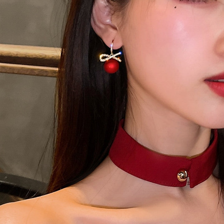 Une femme avec des lèvres rouges et un collier rouge portant des boucles d'oreilles pendantes en forme de boule de Noël rouge avec un nœud orné de strass étincelants. Parfaites pour illuminer vos tenues festives.