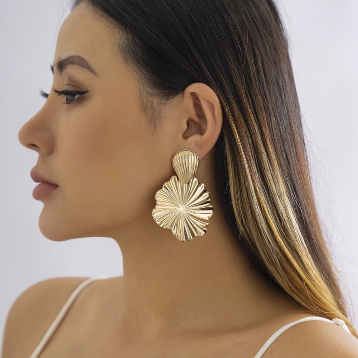 Une femme porte des boucles d'oreille pendantes en métal doré plissé avec un coquillage et une fleur. Un accessoire élégant pour évoquer la beauté intemporelle de la mer. Parfait pour une journée en bord de mer ou une soirée estivale.