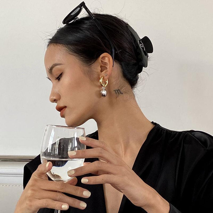 Une femme tenant un verre de vin, avec des boucles d'oreille bicolores en forme de créole et un pendentif en forme de cœur. Bijou contemporain pour femme moderne et audacieuse.