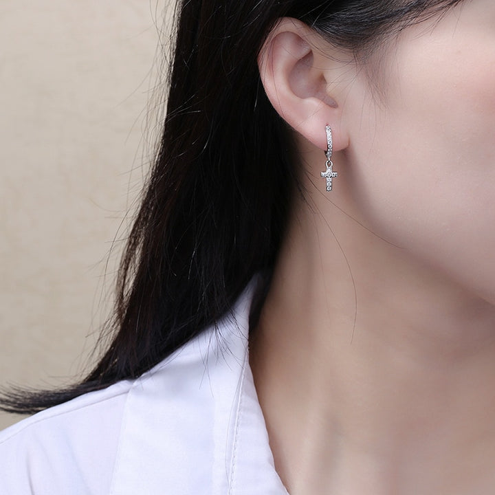 Une femme portant des boucles d'oreille en forme d'anneau avec une croix pendante ornée de strass. Bijou en argent 925, disponible en argent plaqué or et en argent plaqué or rose. Ajoutez de la lumière et de la spiritualité à votre look avec ces boucles d'oreille pendantes.