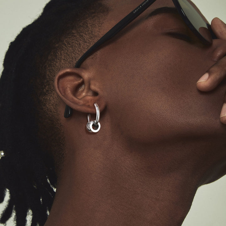 Un homme portant des lunettes de soleil et une boucle d'oreille en acier inoxydable argent avec un motif tressé vieilli. Boucle d'oreille créole anneau stressé viking pour homme, inspirée des guerriers vikings. Allure celte et robuste. Dimensions : 19 x 2,5 mm.