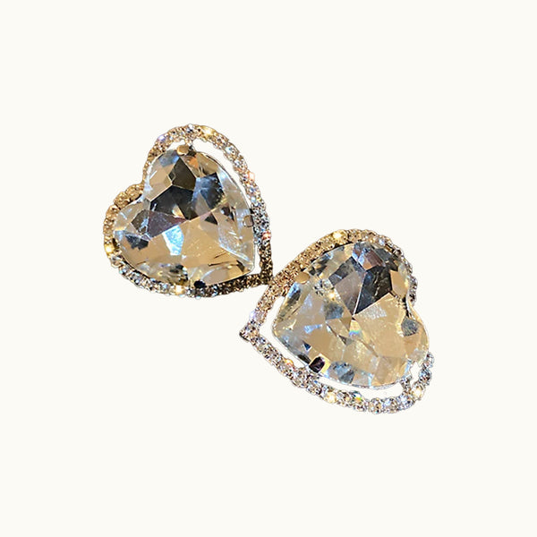 Une paire de boucles d'oreilles sans trou est sur un fond beige. Il y a un coeur en cristal avec un tour en petits diamants ajourés. 