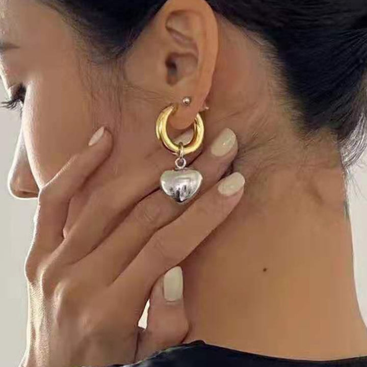 Une femme avec la main sur l'oreille, portant des boucles d'oreille créoles bicolores en or et argent avec un cœur pendu. Parfait pour la femme moderne et audacieuse.