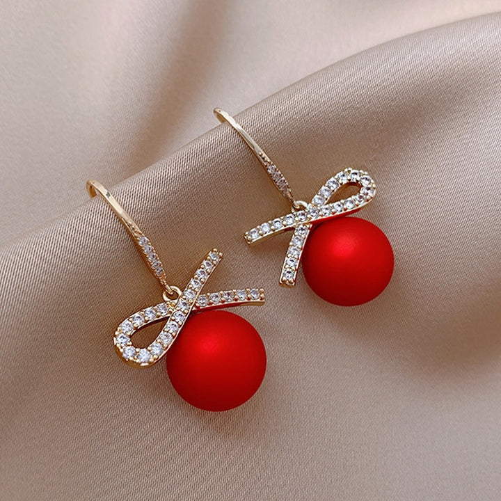 Une paire de boucles d'oreille pendantes en forme de boule de Noël rouge avec un nœud orné de strass étincelants. Parfaites pour illuminer vos tenues festives.