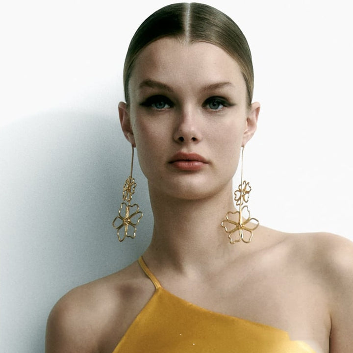 Une femme porte une robe dorée et des boucles d'oreille pendantes en chaîne avec des fleurs métalliques. Un design élégant et moderne pour une soirée spéciale.