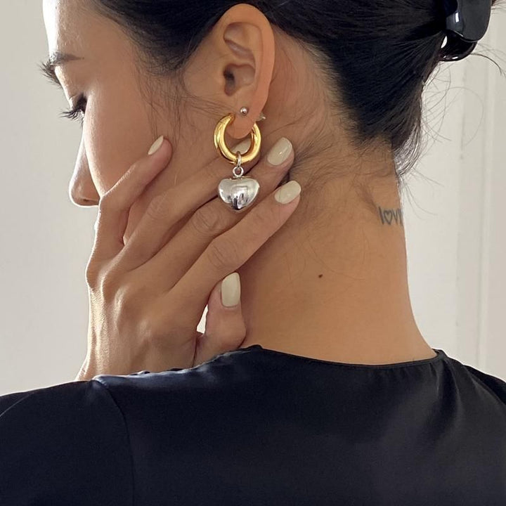 Une femme avec la main sur l'oreille, portant des boucles d'oreille bicolores en forme de créole avec un cœur pendu. Bijou contemporain pour femme moderne et audacieuse.